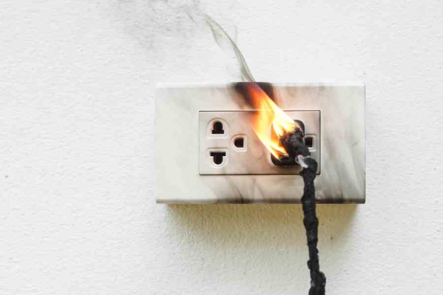 A close-up of an electrical outlet with a plug inserted, showing flames and charring around the outlet.